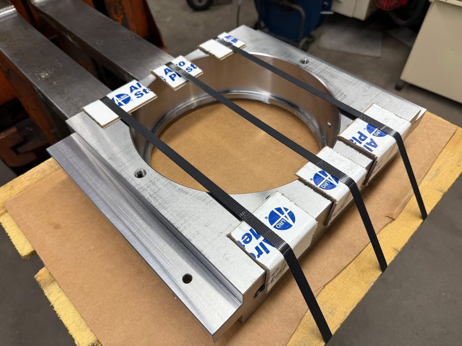 A heavy metal industrial part with a large circular hole, secured with black straps and white protective corner blocks.