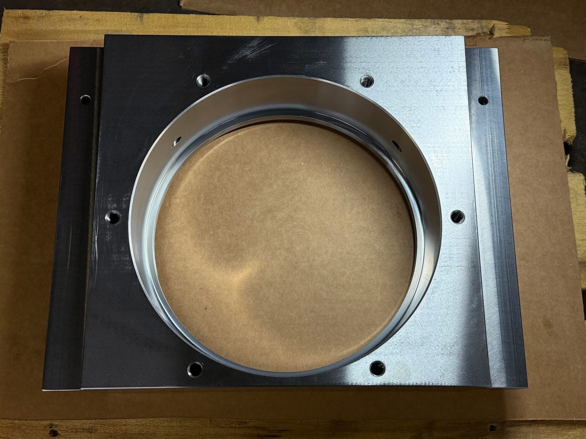 A square, silver-colored metal plate featuring a large central circular opening and several mounting holes on the edges.