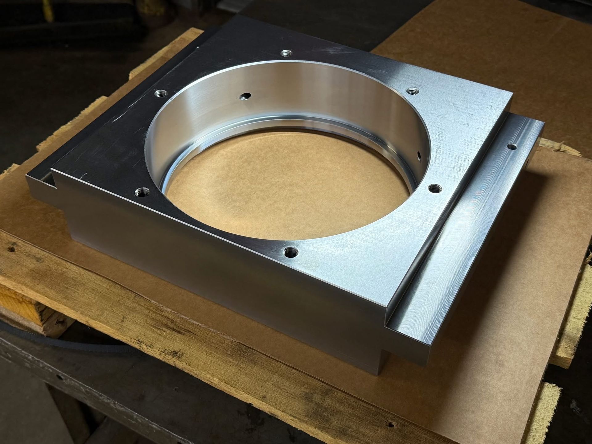 A machined aluminum block with a large circular cutout and several screw holes, resting on a wooden board.