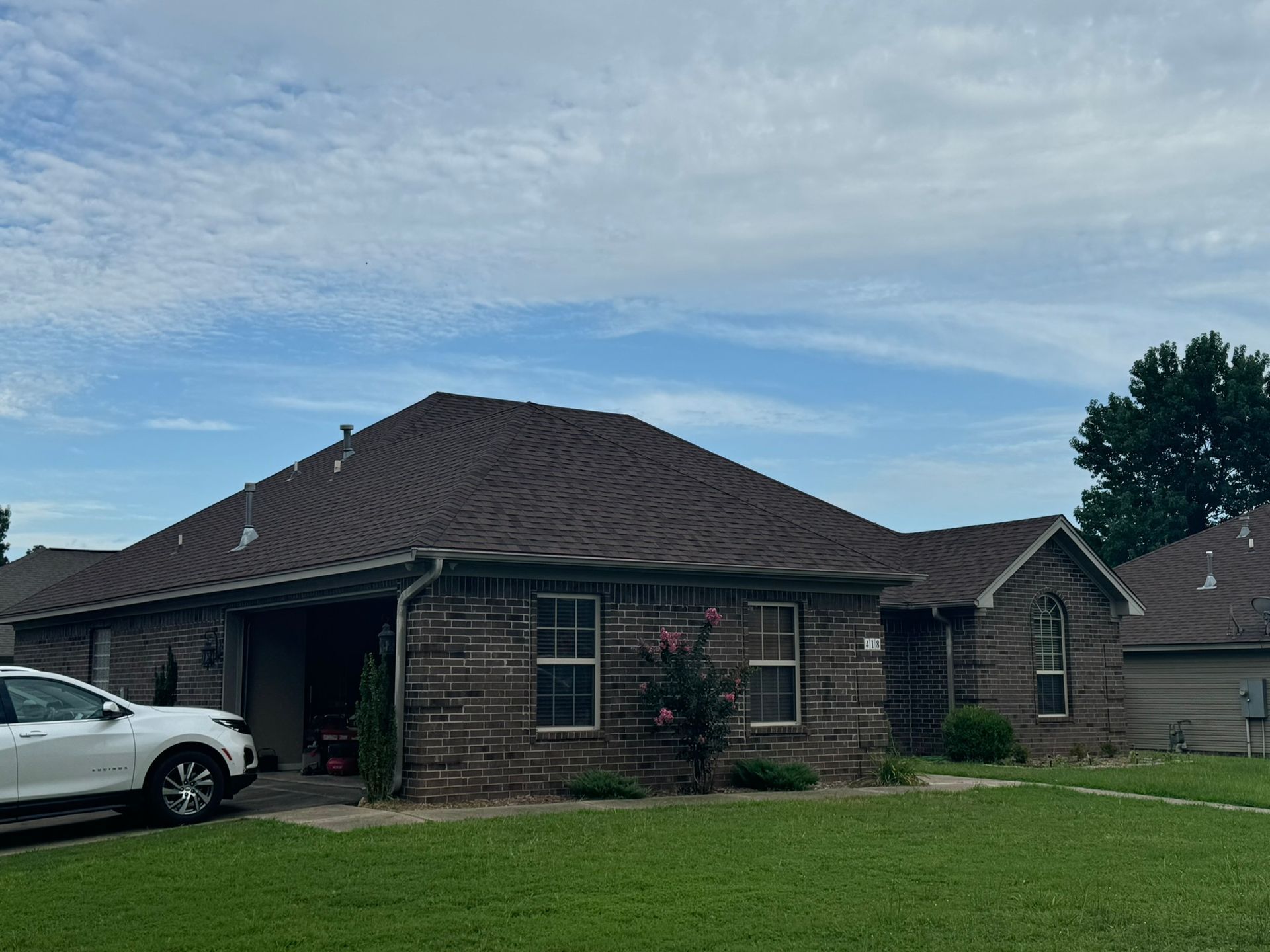 Brown brick house with dark roof, white car parked in front, green lawn, cloudy blue sky.
