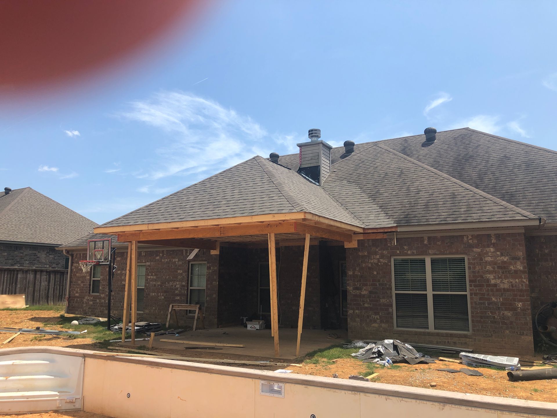 A brick house with a wooden patio under construction, a pool in front, and a blue sky.
