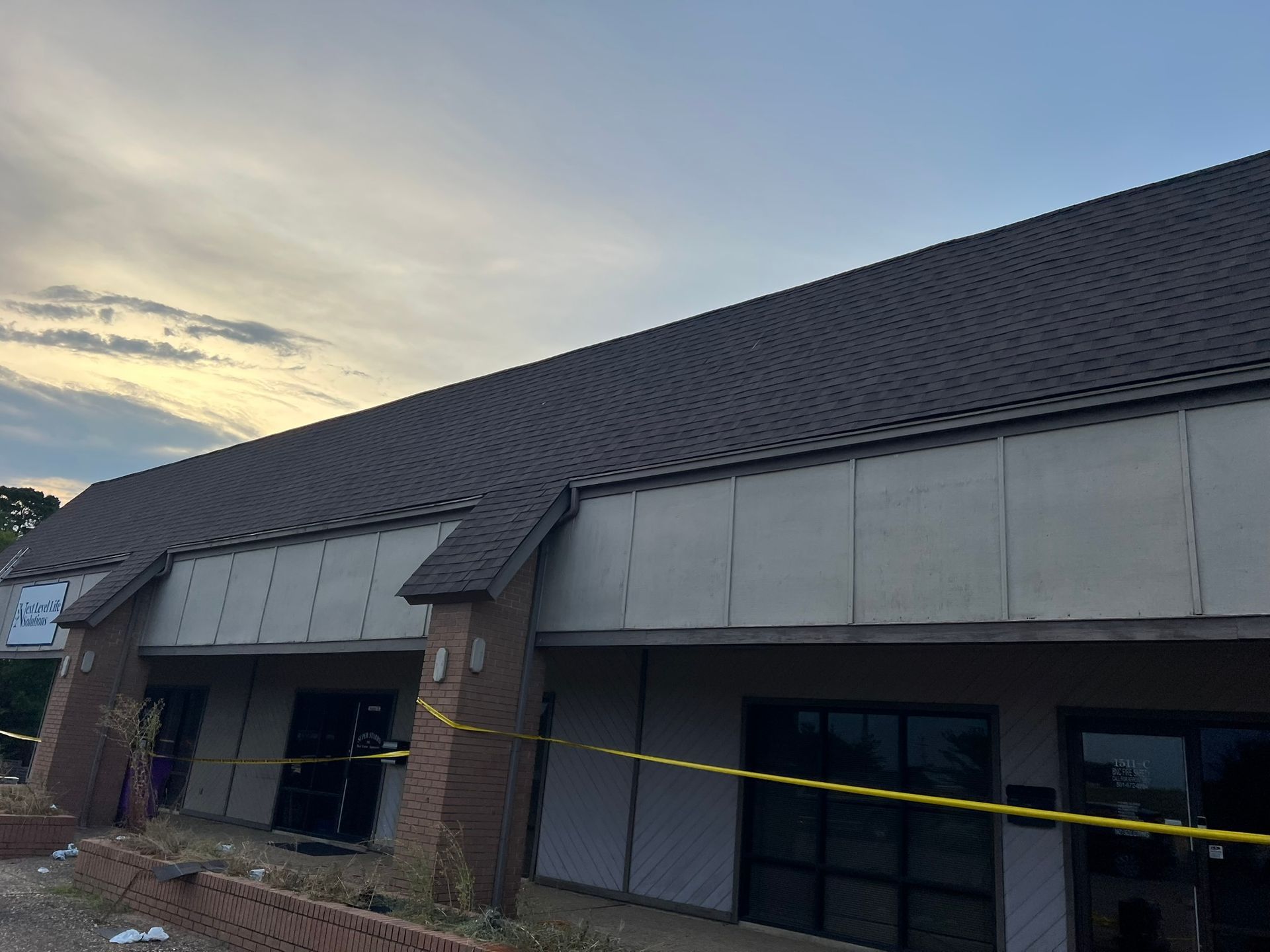 Building exterior with dark roof and beige siding under a cloudy sky. Yellow caution tape present.