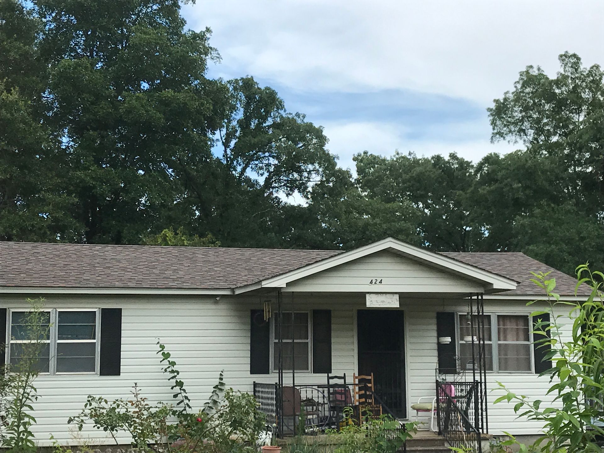 White house with dark shutters, porch, and a brown roof. Trees in background, cloudy sky.