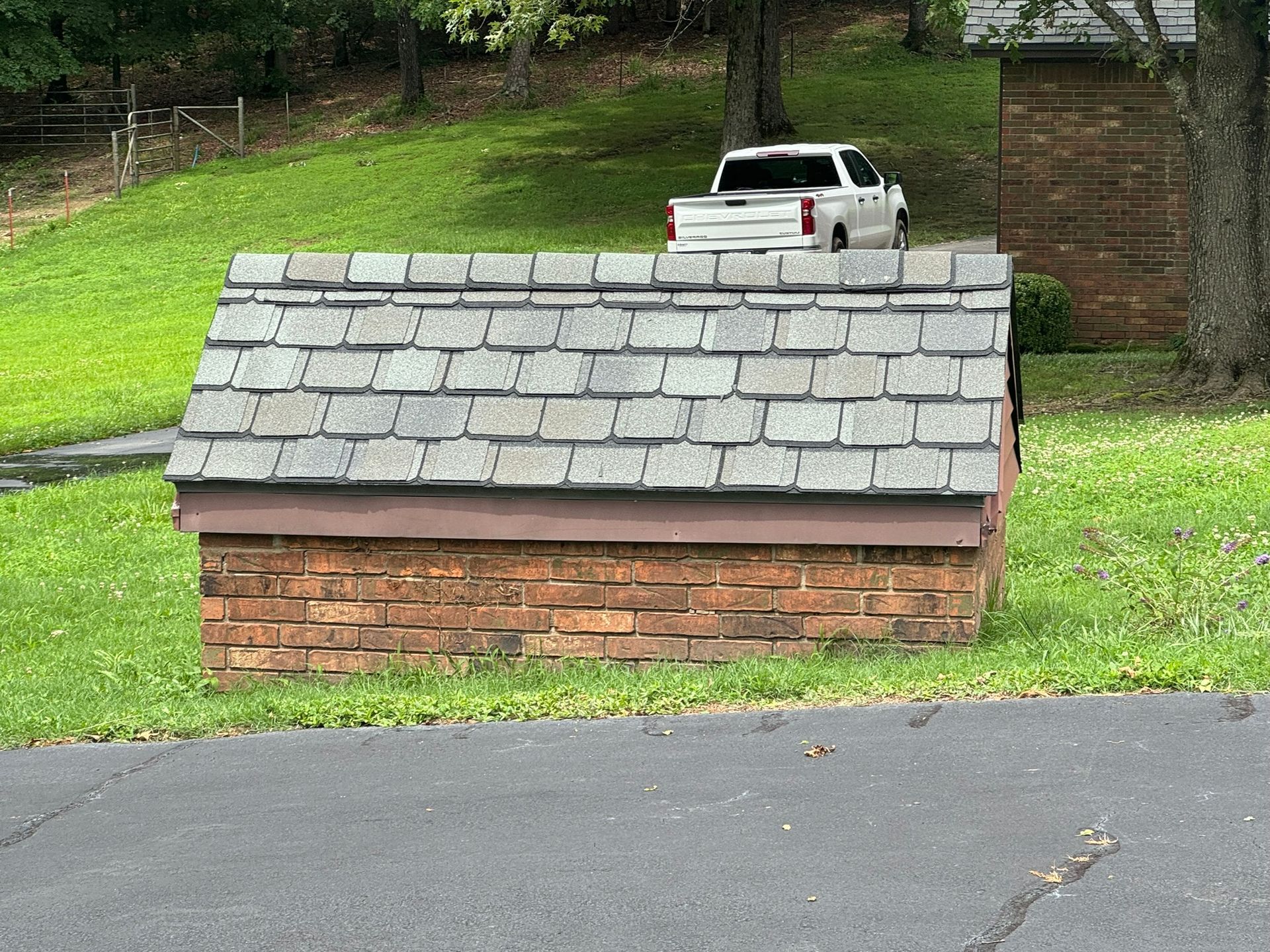 Brick structure with a shingle roof in a grassy area, with a white truck in the background.