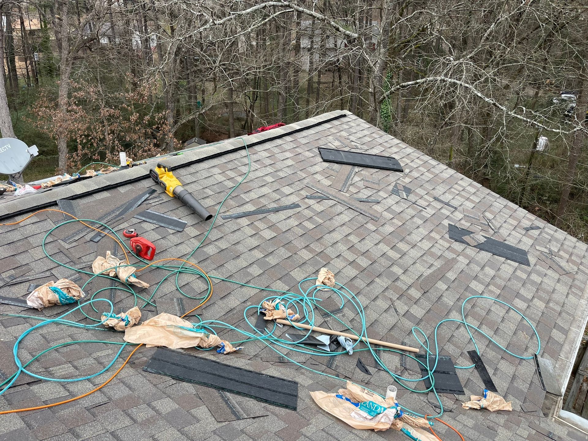 Rooftop with scattered debris, tools, and replacement shingles; trees in background.