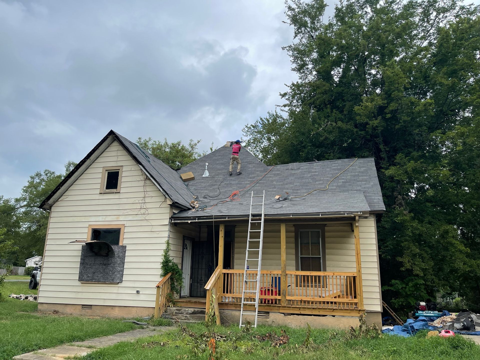 Man on a roof replacing shingles of a weathered, two-story house. Ladder and overcast sky visible.