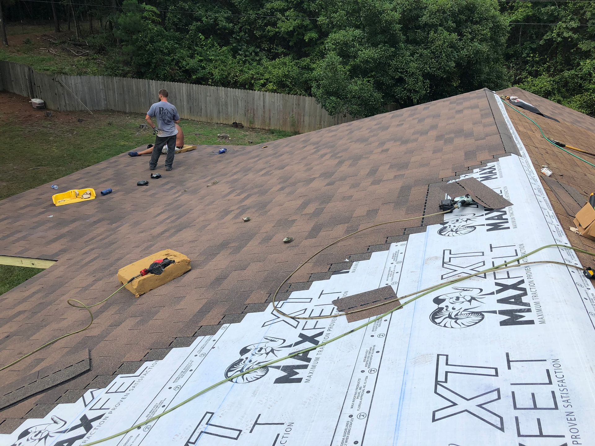 A person installing new shingles on a roof, tools visible. New roofing material and trees in the background.