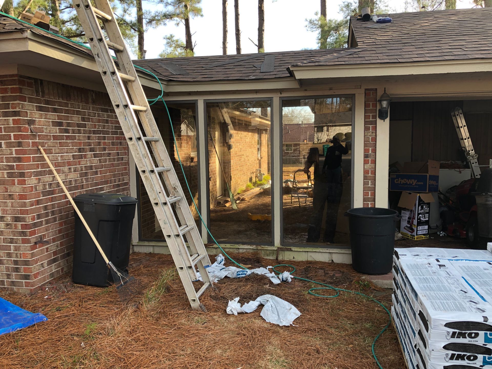 Ladder leaning against a house with a person visible inside. Roofing materials and trash cans are nearby.