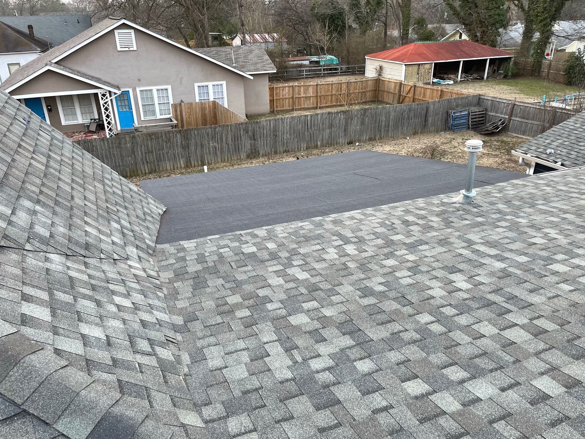 View from a roof of surrounding houses with gray shingles, fence, and paved area in the backyard.