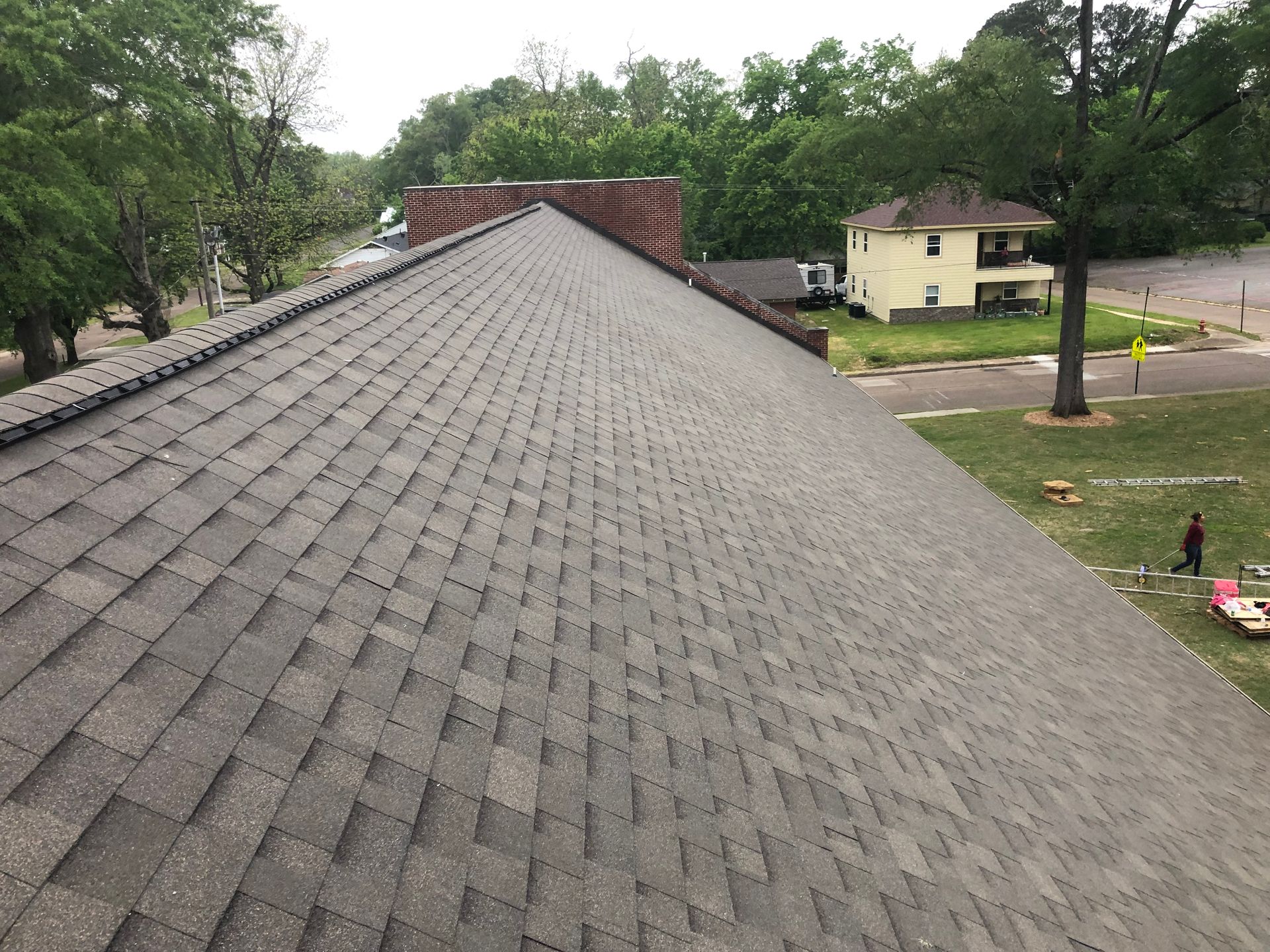 Overhead view of a gray shingled roof with a brick chimney, trees, and houses in the background.