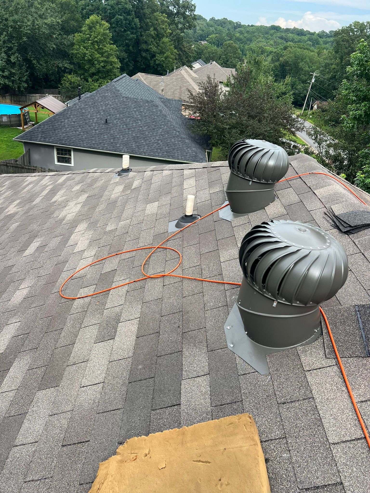 View of a roof with two spinning vents, with houses and trees in the background. An orange safety rope is visible.