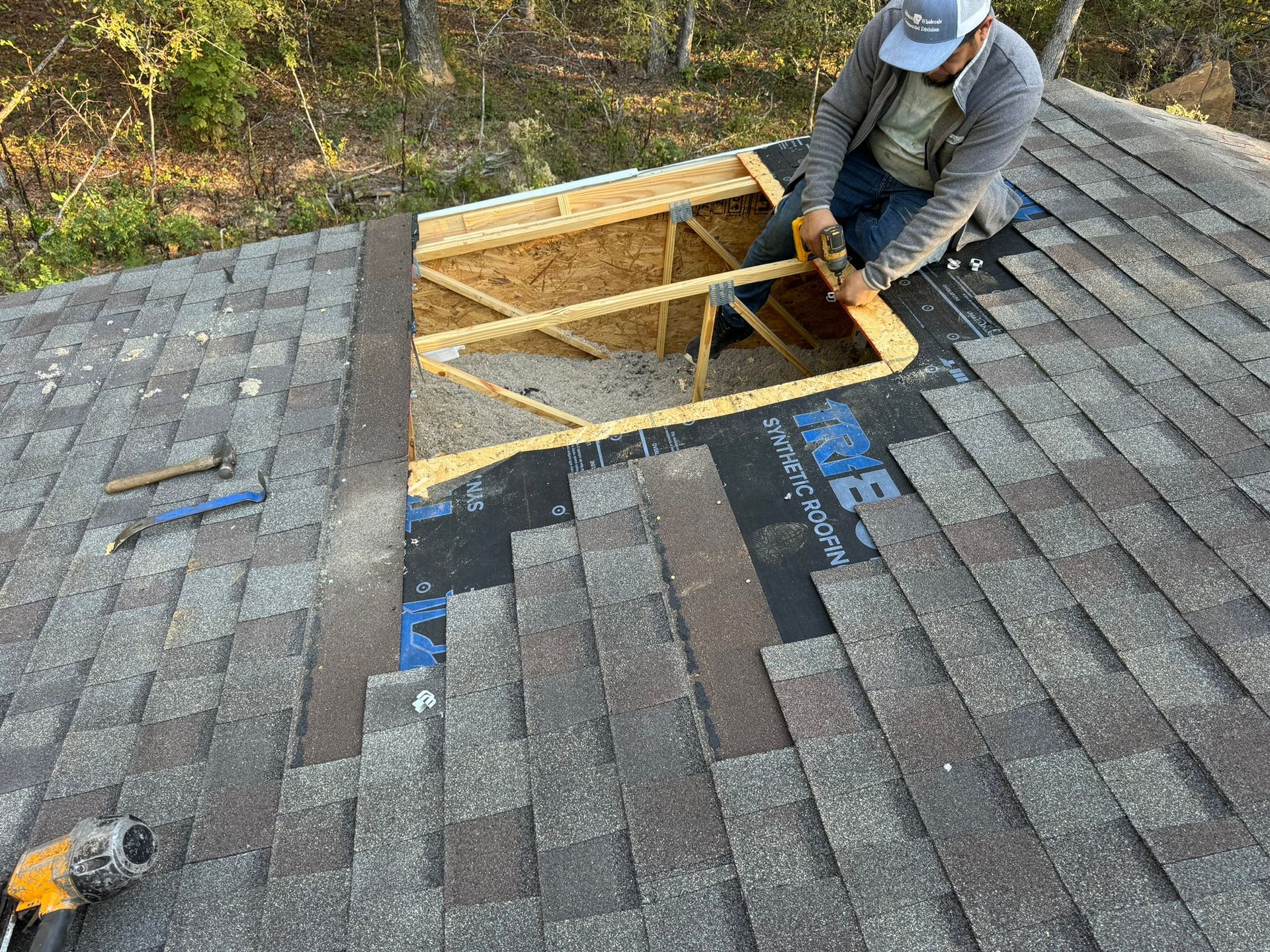 Man on roof, cutting wood around an opening. Shingles and roof underlayment visible.