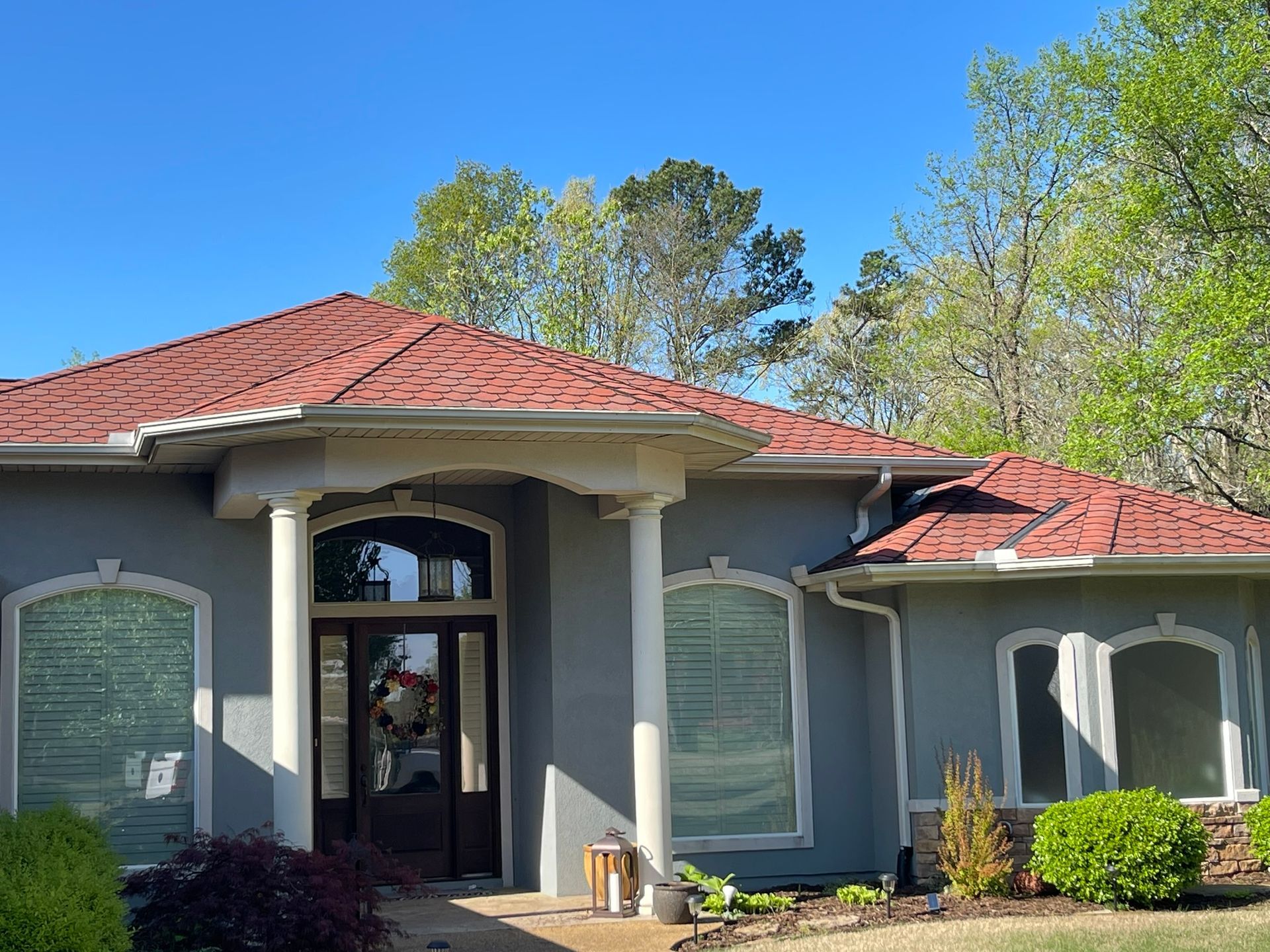 Gray stucco house with red tile roof, white columns, and lush greenery under a clear blue sky.