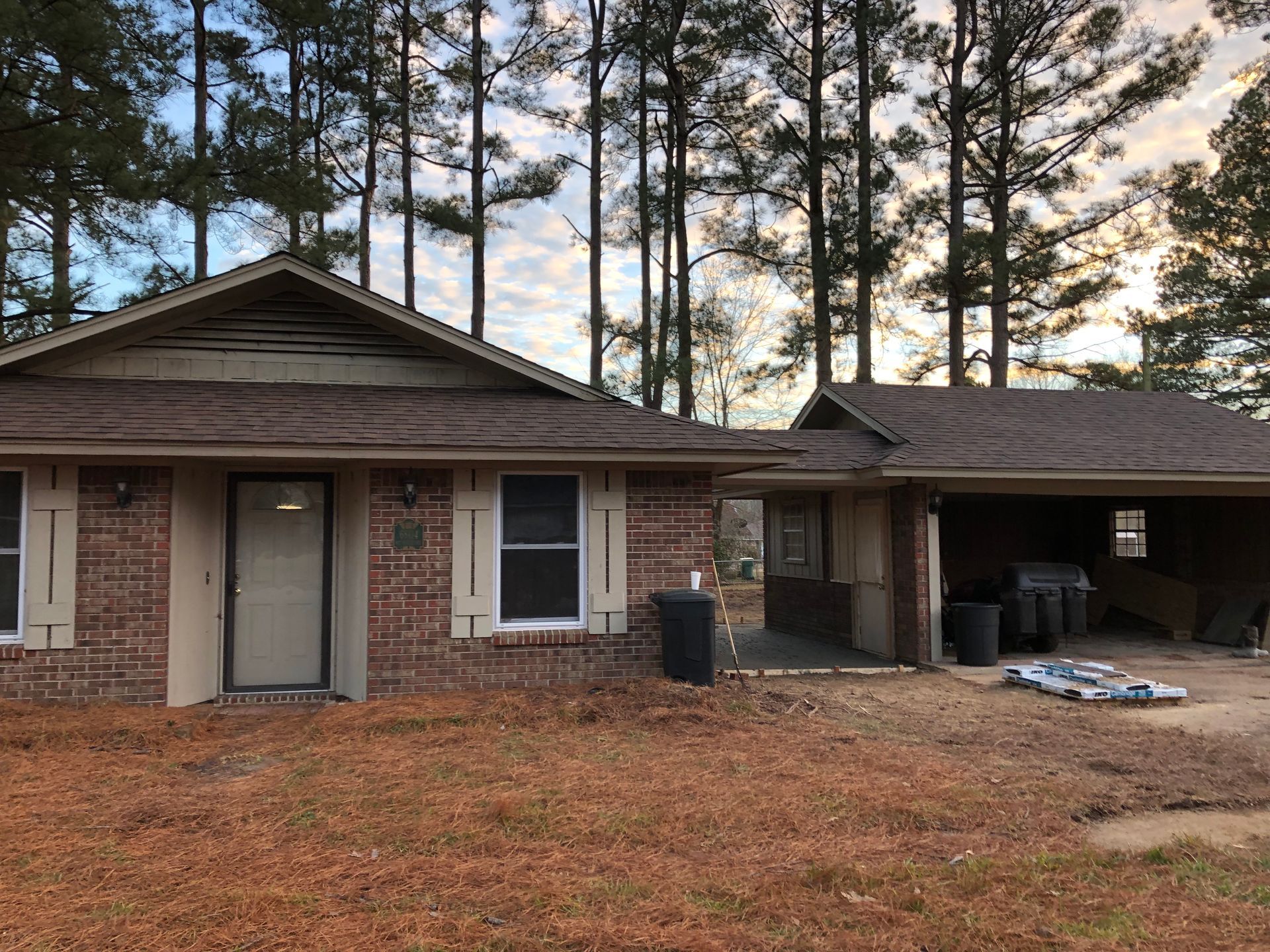Brick house with carport; brown roof, door, and windows. Dead grass. Tall trees in background.
