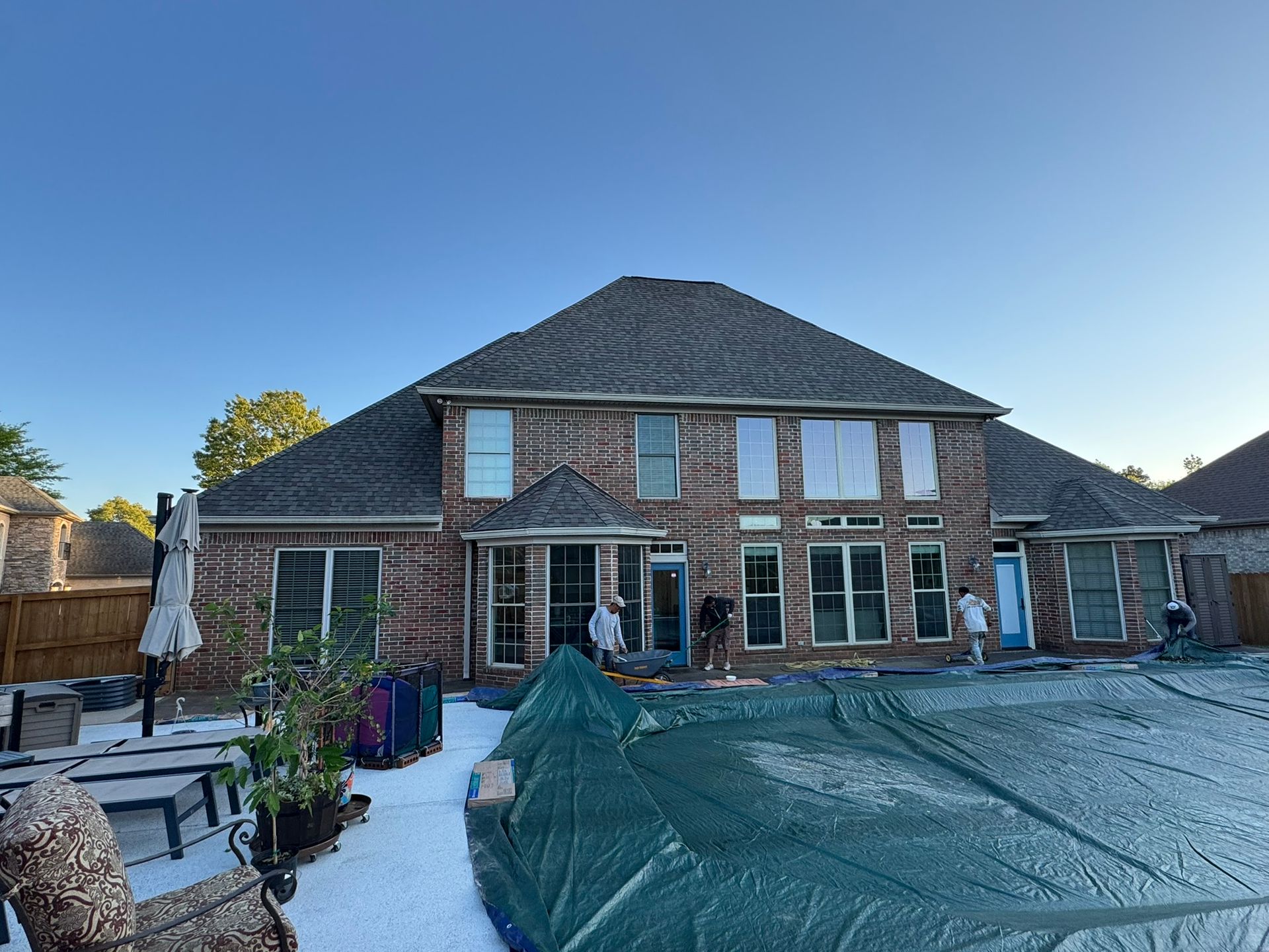 Back view of a brick house with a dark roof, pool covered with a tarp, and people standing near the doors.