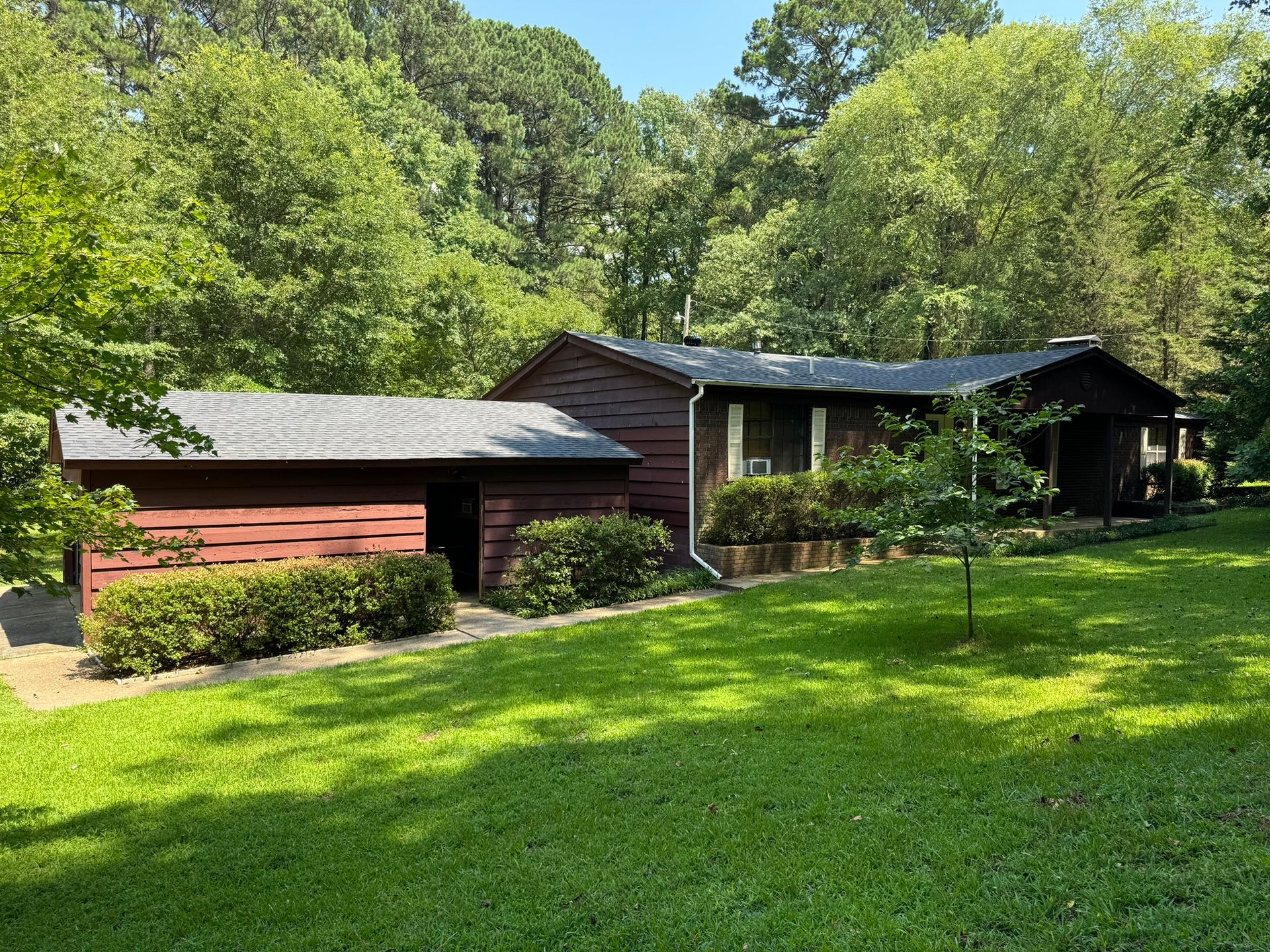 A single-story brown house with a detached garage sits on a green lawn surrounded by trees.