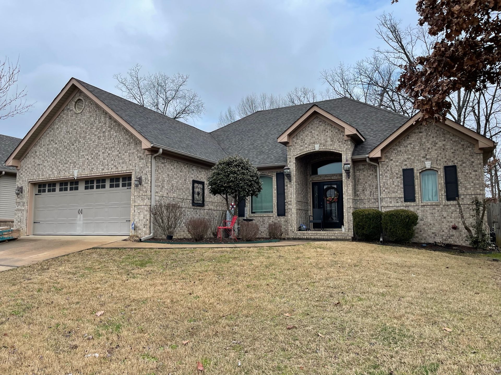 Brick house with a gray roof and a garage, set on a brown, grassy lawn under a cloudy sky.