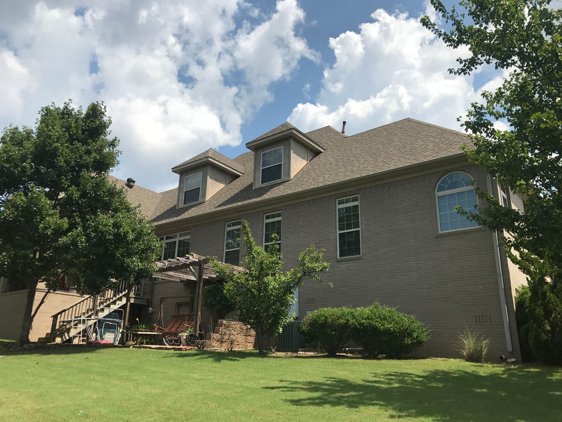 Tan-colored house with dormer windows, surrounded by green grass and trees, against a cloudy sky.