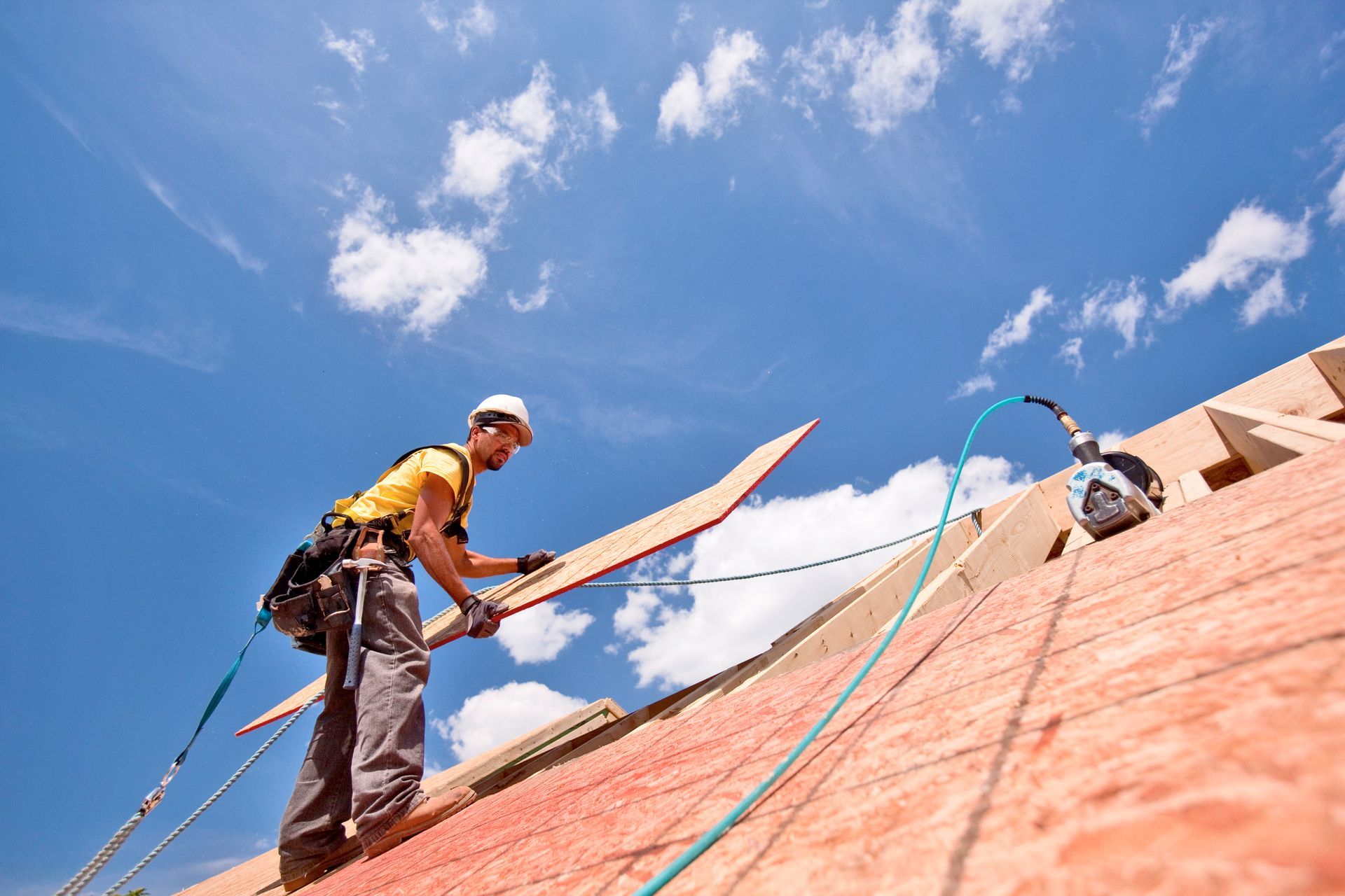 Roofer wearing a harness placing a wood plank on a roof under a blue sky with clouds.