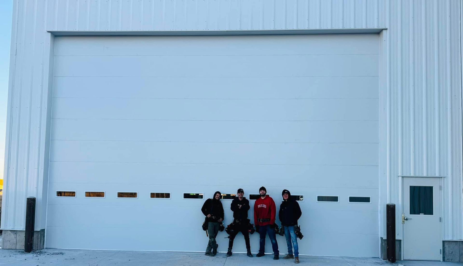 Four people stand in front of a large, closed white garage door; building exterior.