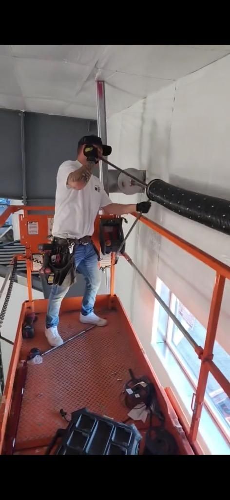 Man on an orange lift drills into a wall with a long tool. He wears a white shirt, jeans, and a tool belt.