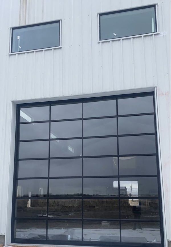 White industrial building with large, divided-pane glass door reflecting the sky and two small rectangular windows above.