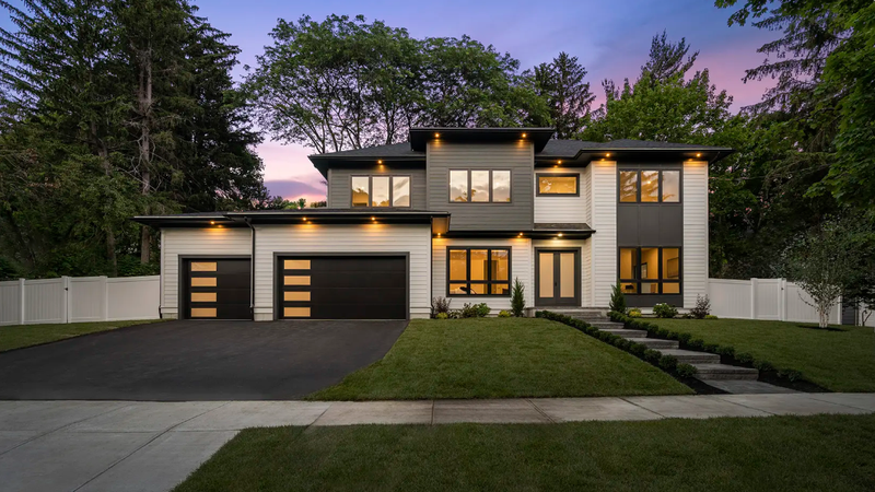 Modern two-story house with black garage doors, gray and white facade, and landscaped front yard at dusk.