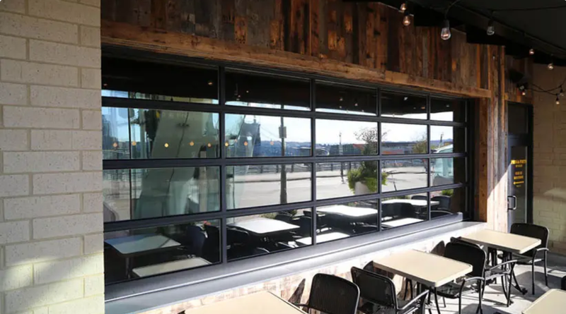 Restaurant with large glass windows and outdoor seating; light-colored brick and wood accents. Tables and chairs visible.