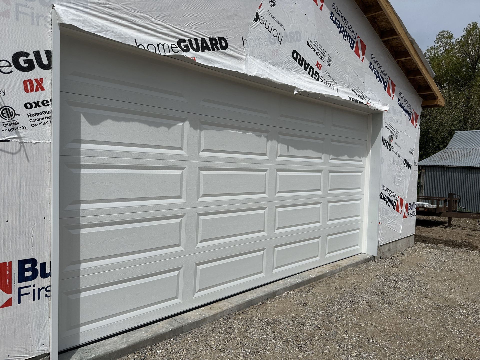 White garage door on a building under construction, covered with home protection wrap, gravel ground.