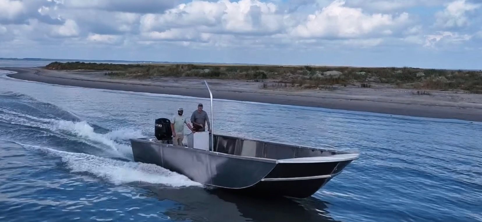 An aluminum boat moves through calm water near a grassy shoreline under a cloudy sky.