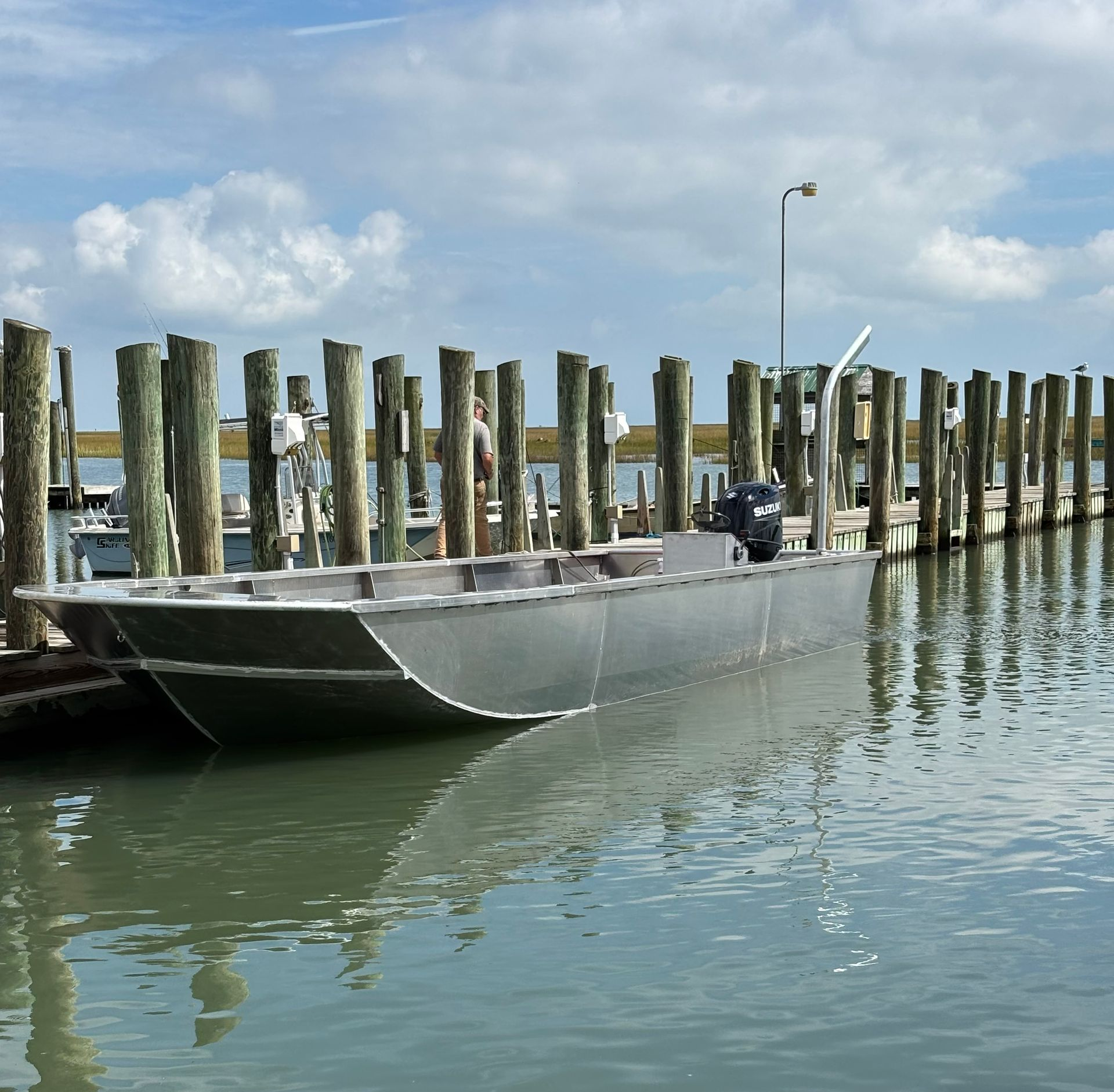 A metal flat-bottomed boat moored at a wooden dock on a calm, cloudy day.