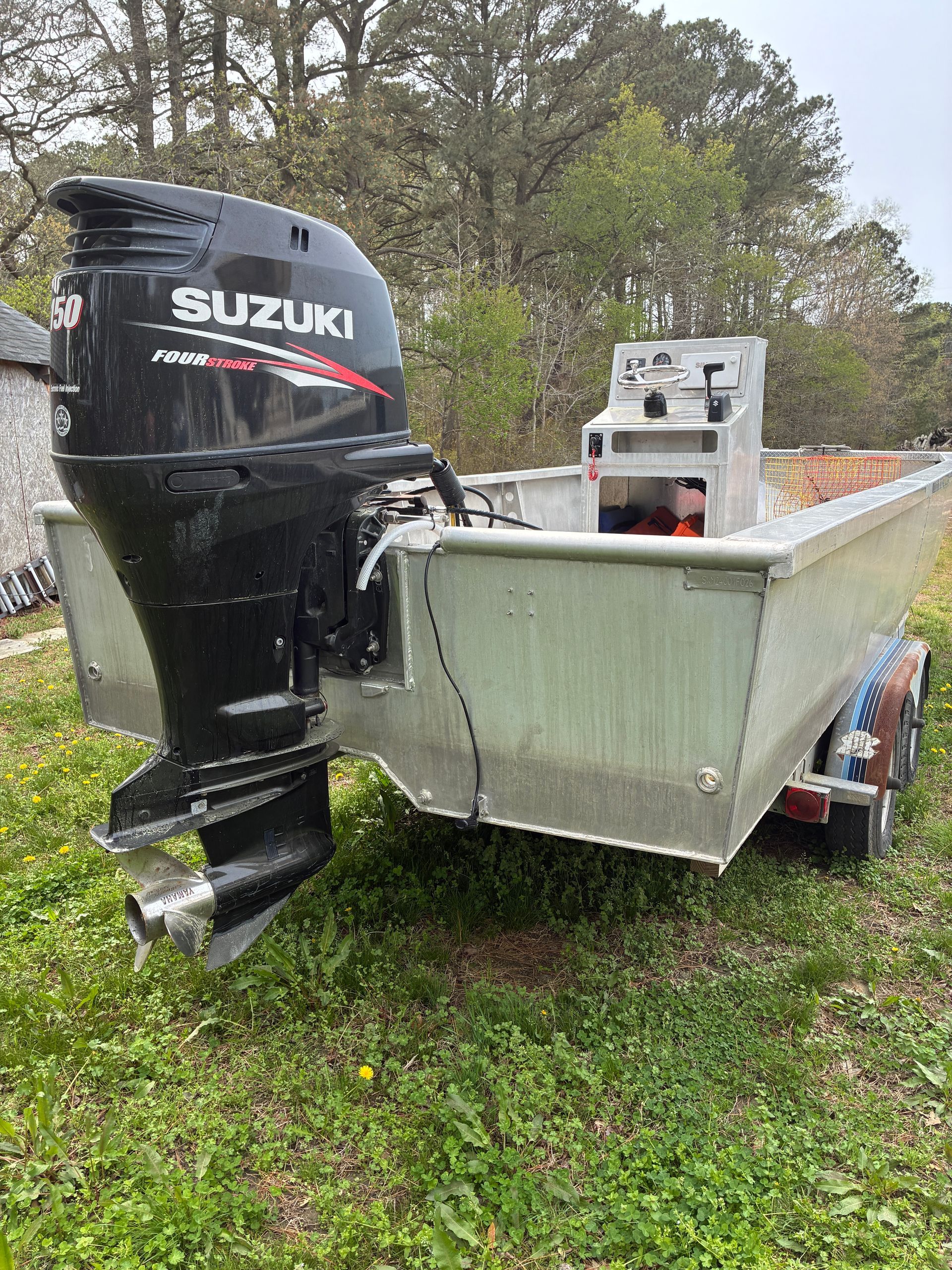 A silver aluminum boat with a Suzuki outboard motor, mounted on a trailer in a grassy, wooded area.