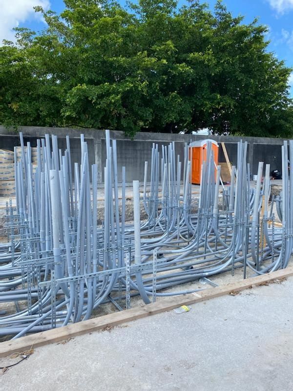 Gray metal conduits piled at a construction site, with an orange portable toilet and green trees in the background.