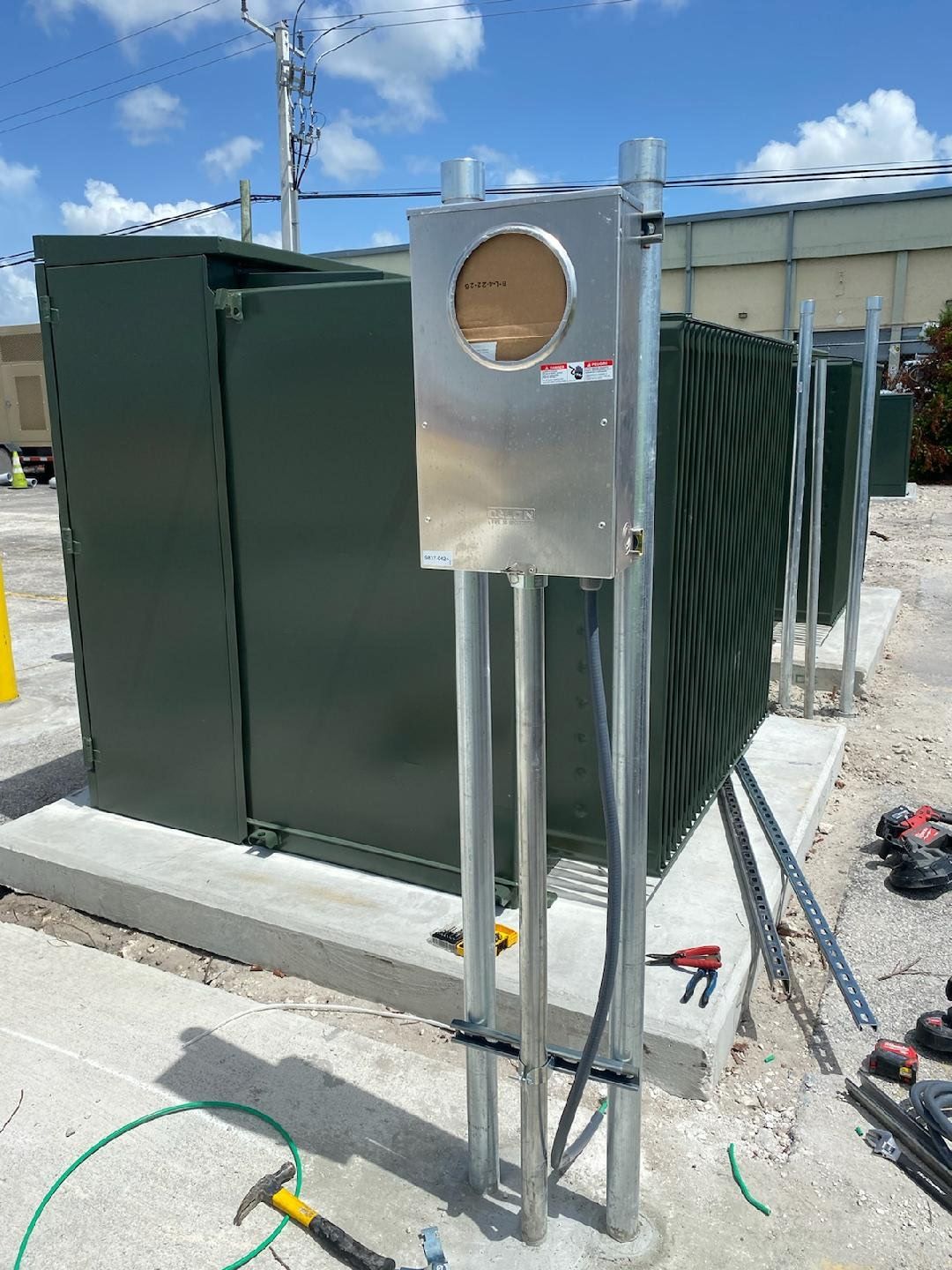 Metal electrical meter box with conduit pipes, set on a concrete pad, with large green electrical equipment in the background.