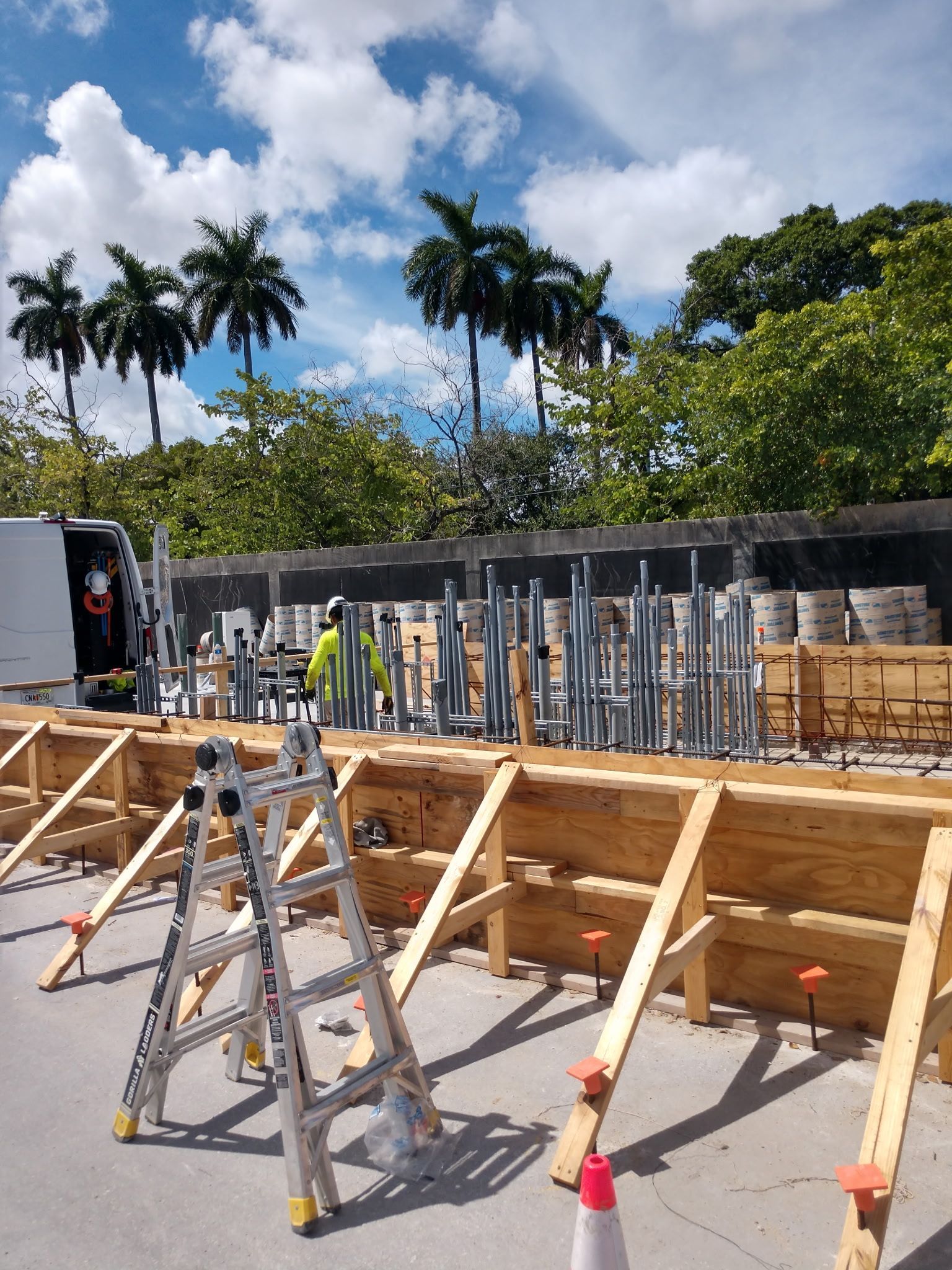 Construction site with wooden forms, metal scaffolding, and workers. Palm trees and blue sky background.