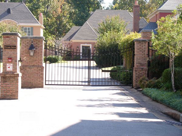 A black wrought iron gate leading into a residential community, flanked by brick pillars and greenery.