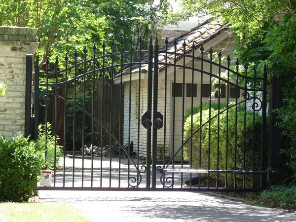 Black wrought-iron gate closed over a paved driveway, leading to a light-colored building.
