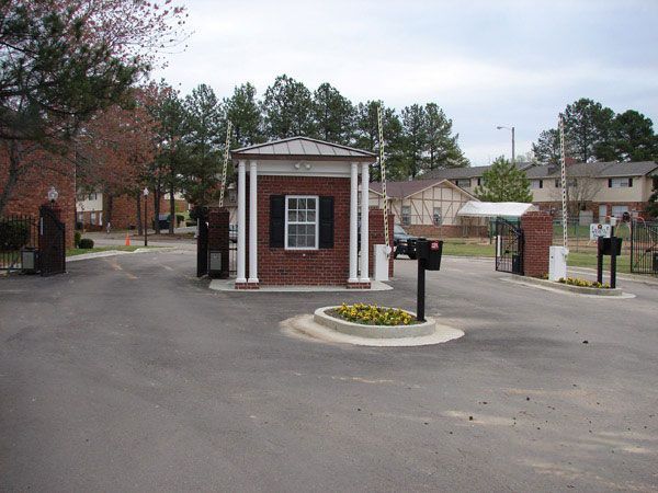 Entrance to a gated community with a guard house in the center, flanked by brick pillars and open gates. Asphalt road with a small flower bed.
