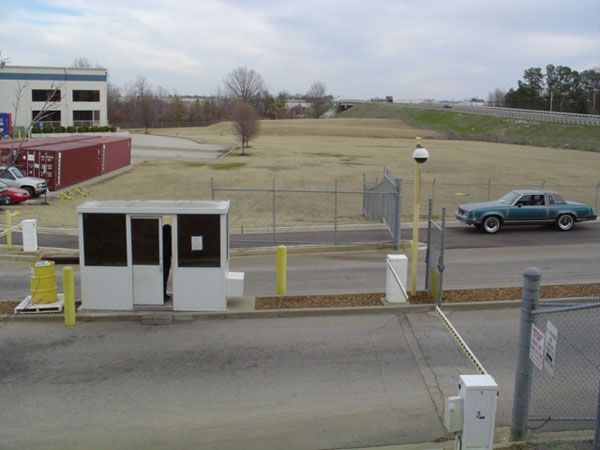 A guard booth at an entrance gate with a teal car driving through; a building and brown containers are visible in the background.