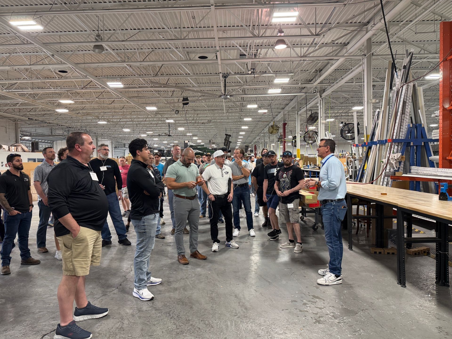 People gathered in a bright warehouse, listening to a man speaking near worktables and equipment.