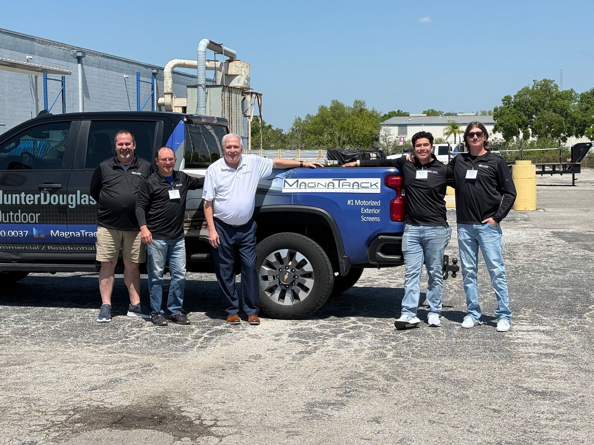 Five people posing beside a blue pickup truck in an industrial lot on a sunny day