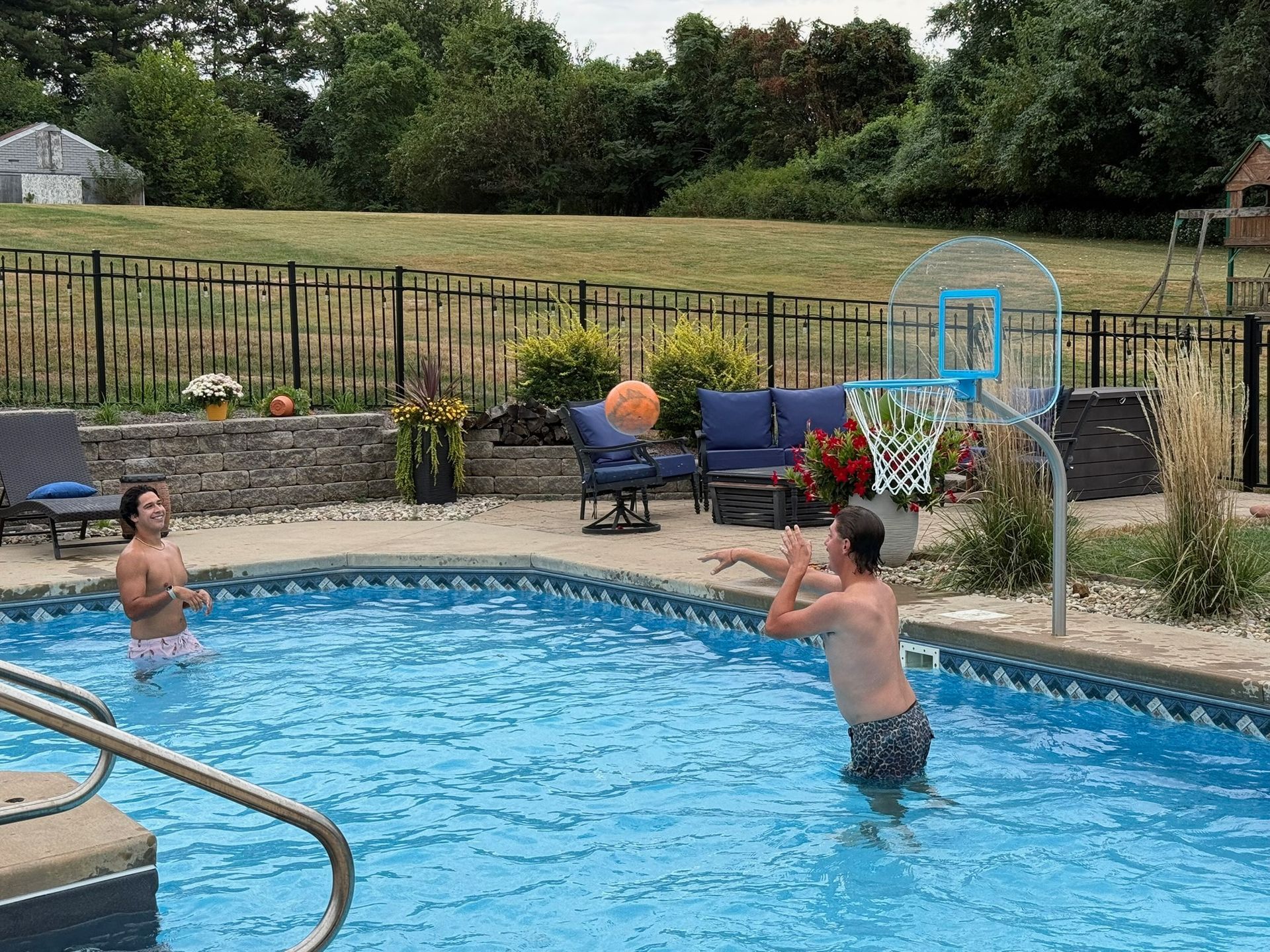 Two people in a backyard pool playing catch near a basketball hoop