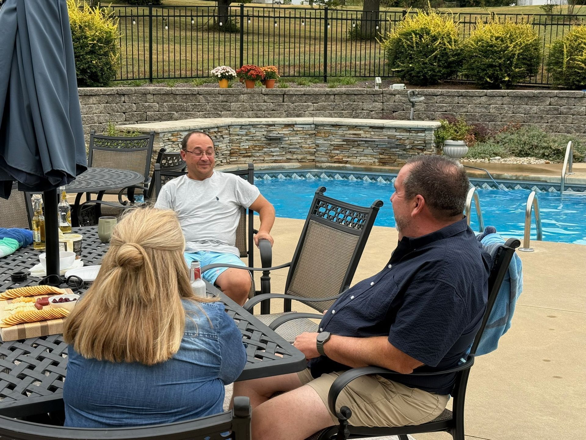 People sitting around a poolside table with chairs, chatting outdoors near a backyard pool.