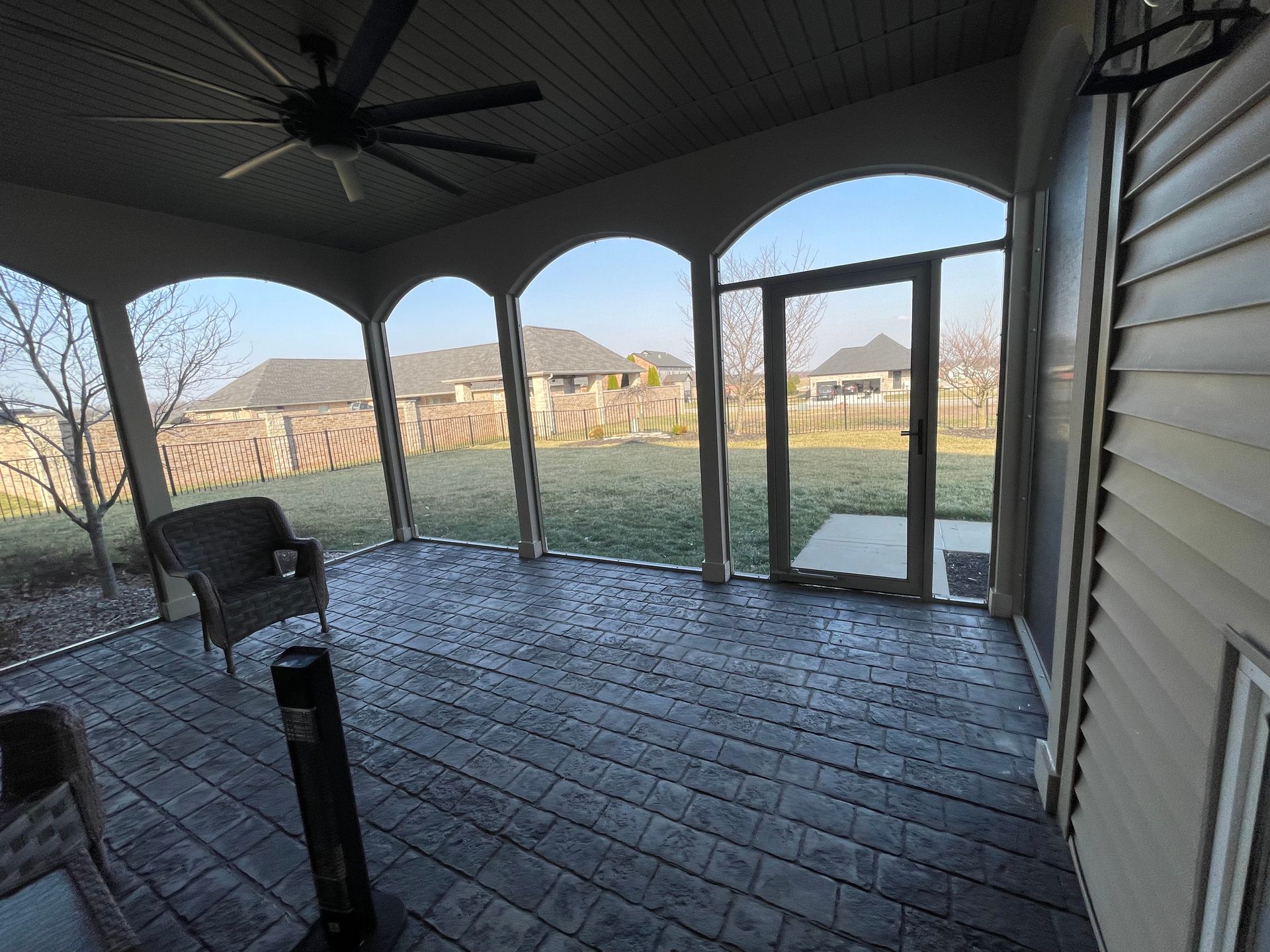 Screened-in porch with arched openings, wicker furniture, and a fan. View of a backyard.