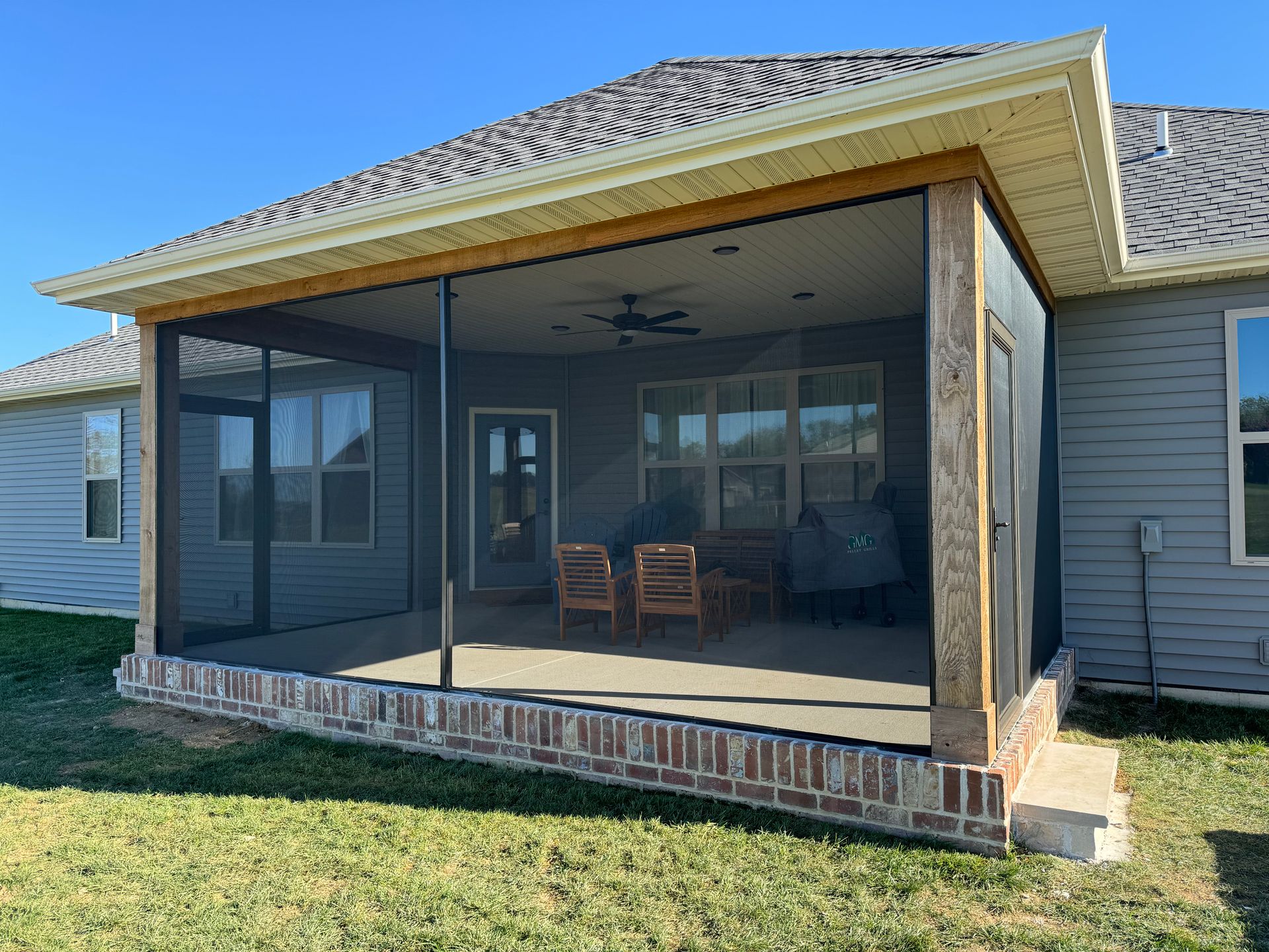 Screened-in porch with brick base, light-colored trim, and outdoor furniture, attached to a house with a blue sky.