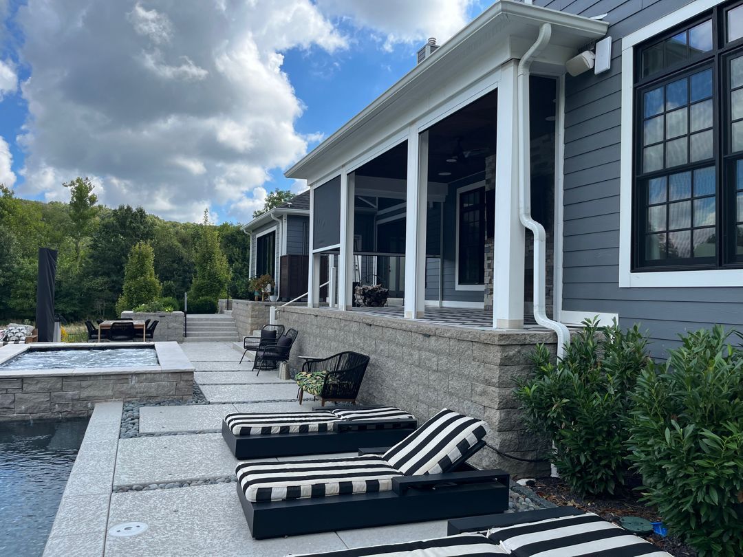 Poolside scene with lounge chairs, outdoor kitchen, and a gray house with blue sky and trees.