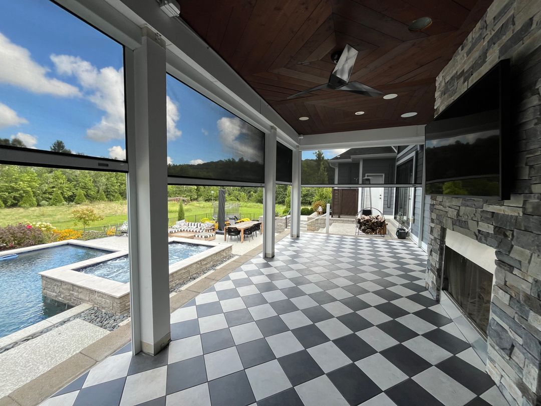 Outdoor patio with pool, fireplace, black and white checkered floor, retractable shades, and open view.
