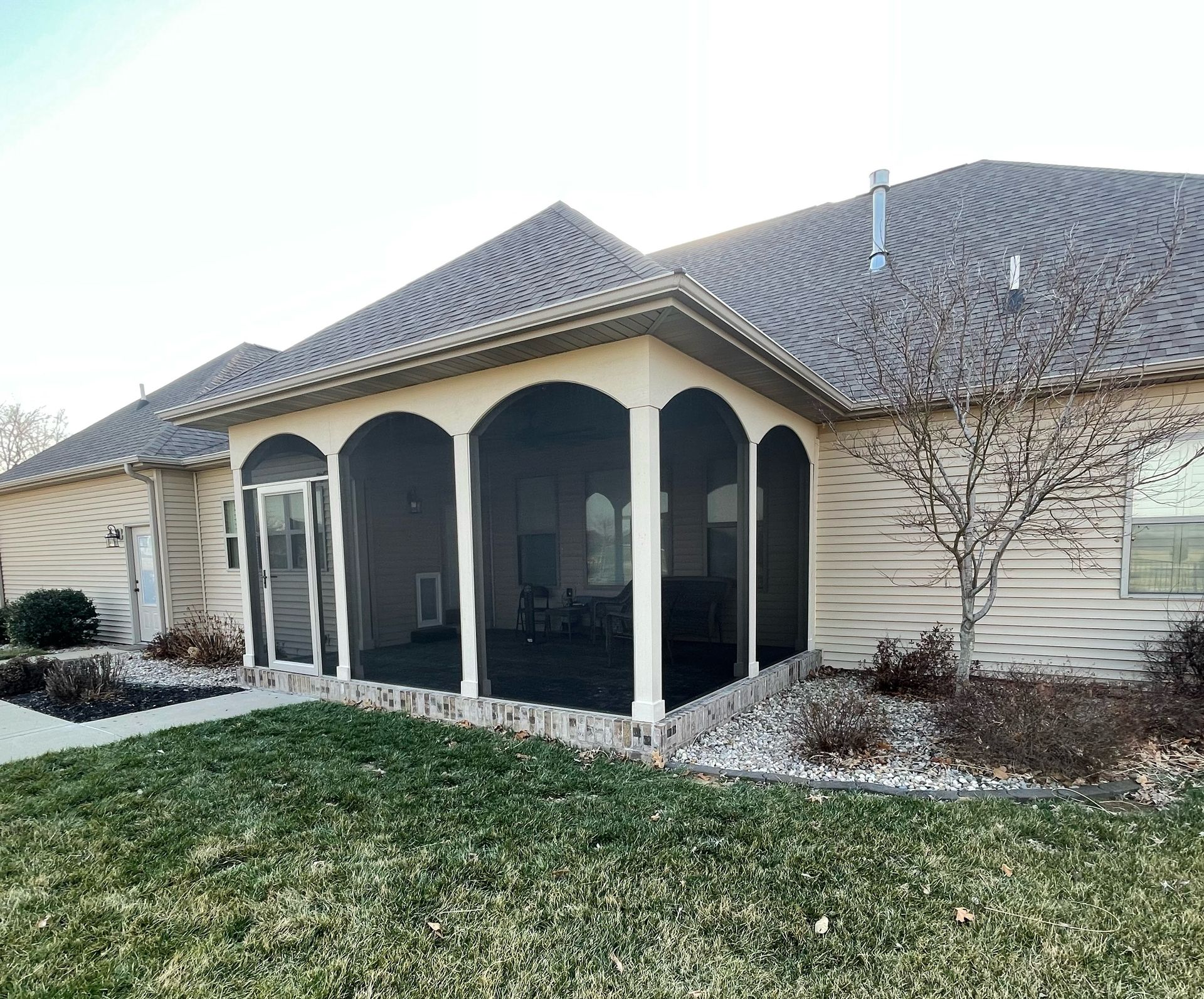 Screened-in porch attached to a house with arched openings and dark screens, on a grassy lawn.
