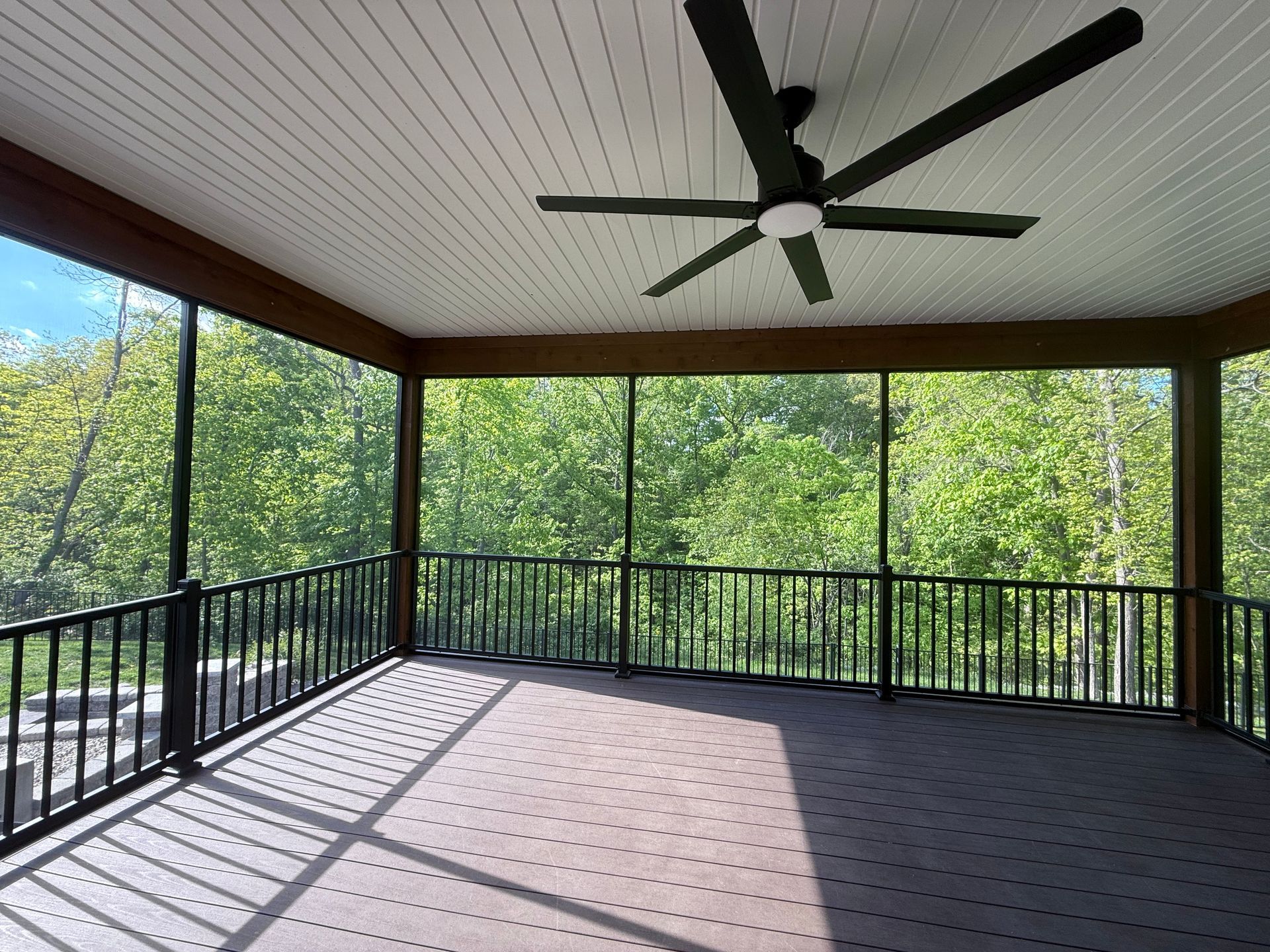 Screened porch with dark railing, composite deck, ceiling fan, and a view of trees.
