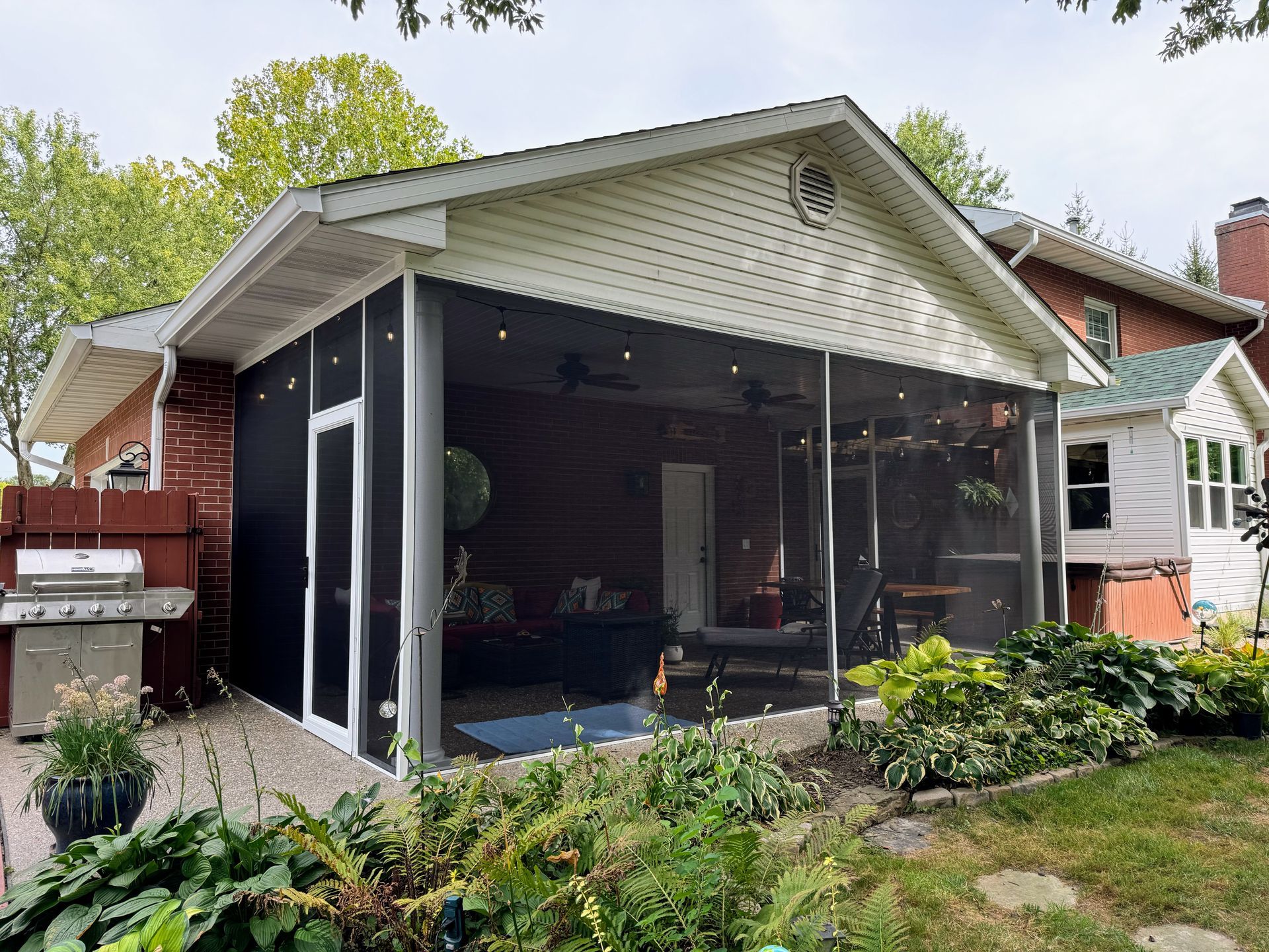 Screened-in porch attached to a brick house with a grill and garden in the yard.